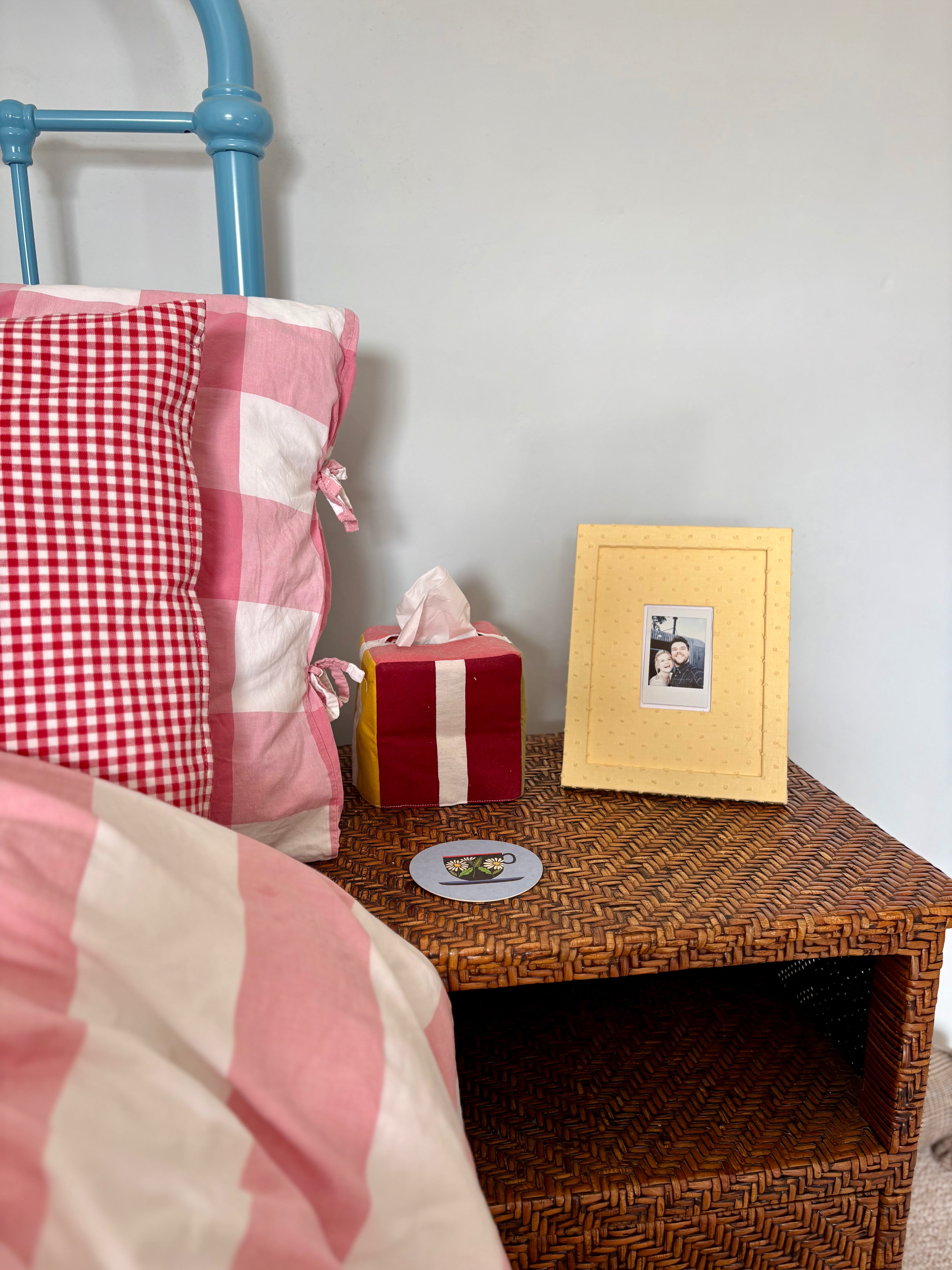 Bed with pink checkered pillow and red blanket, nightstand with tissue box and photo frame.